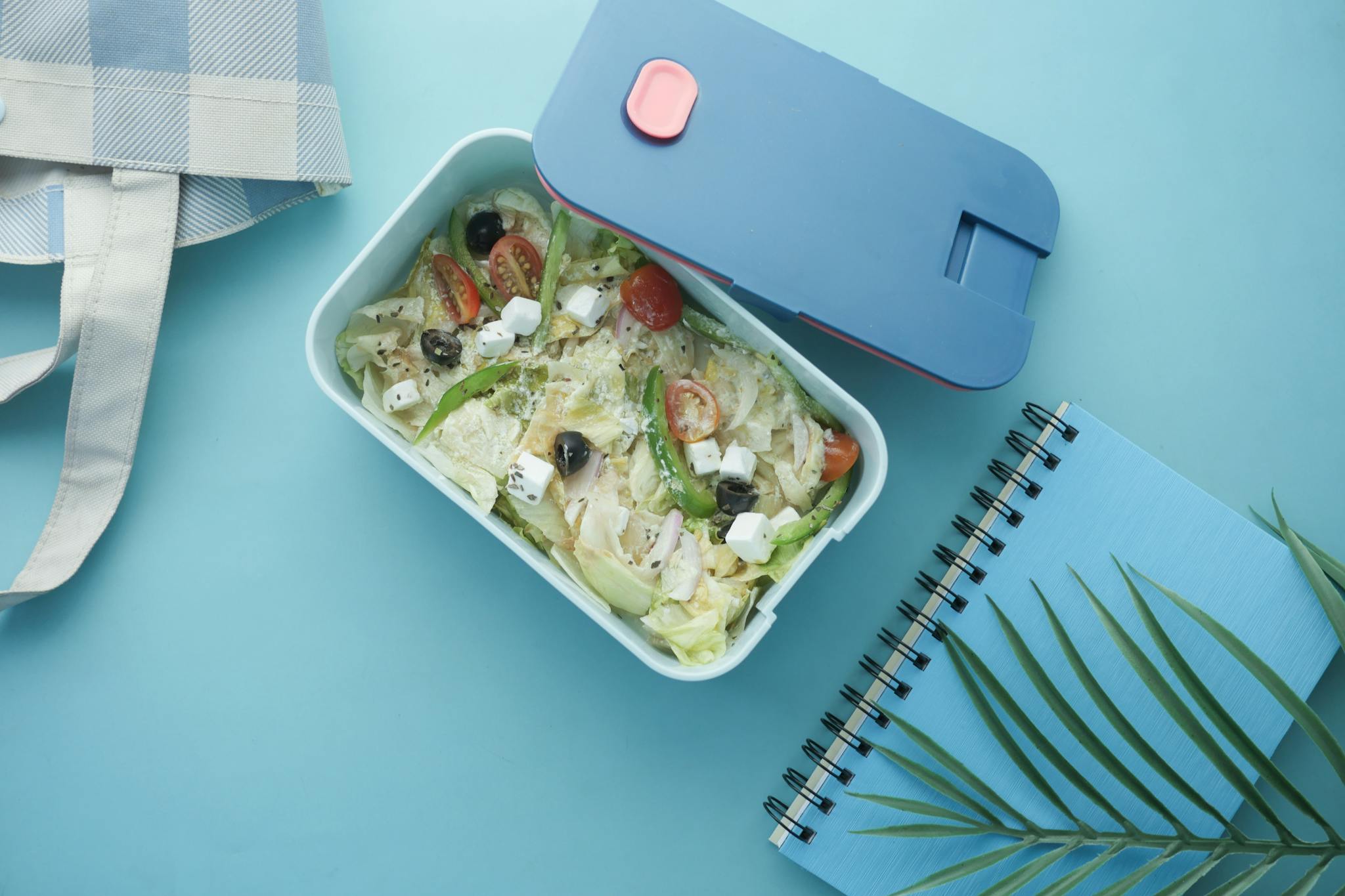 Top view of a nutritious salad lunch box with a notebook on a blue background.
